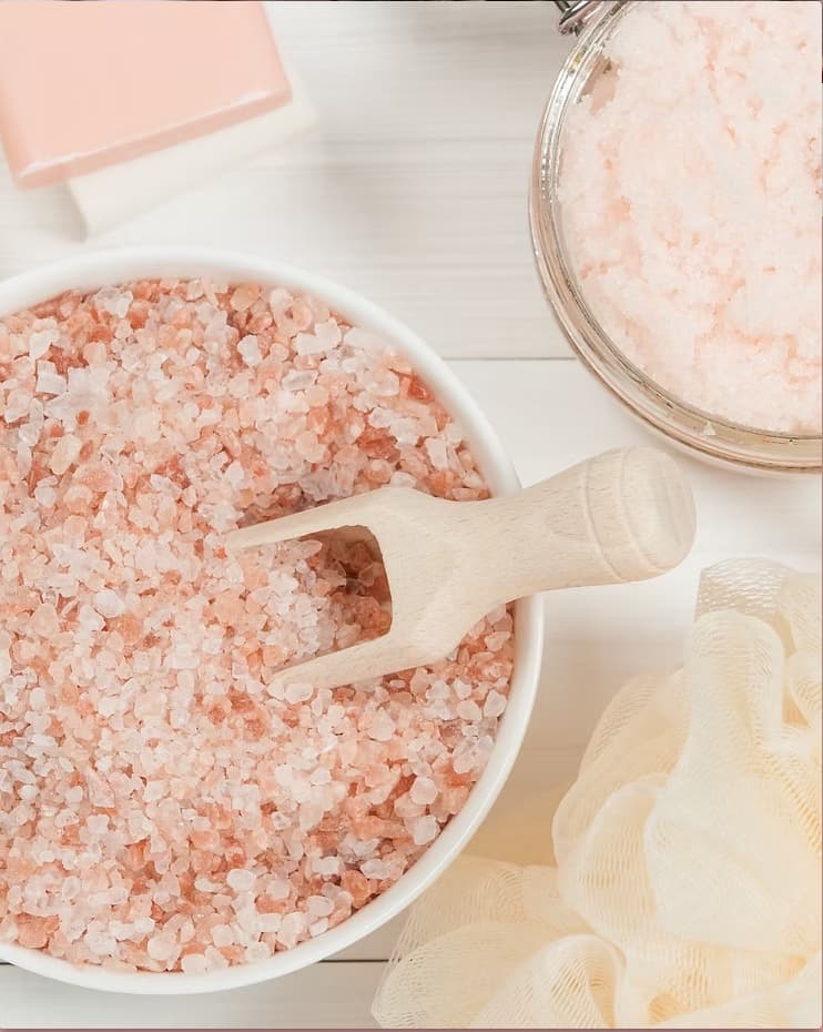 A bowl of pink Himalayan salt scrub with a wooden scoop, beside a soap bar and a soft bath pouf